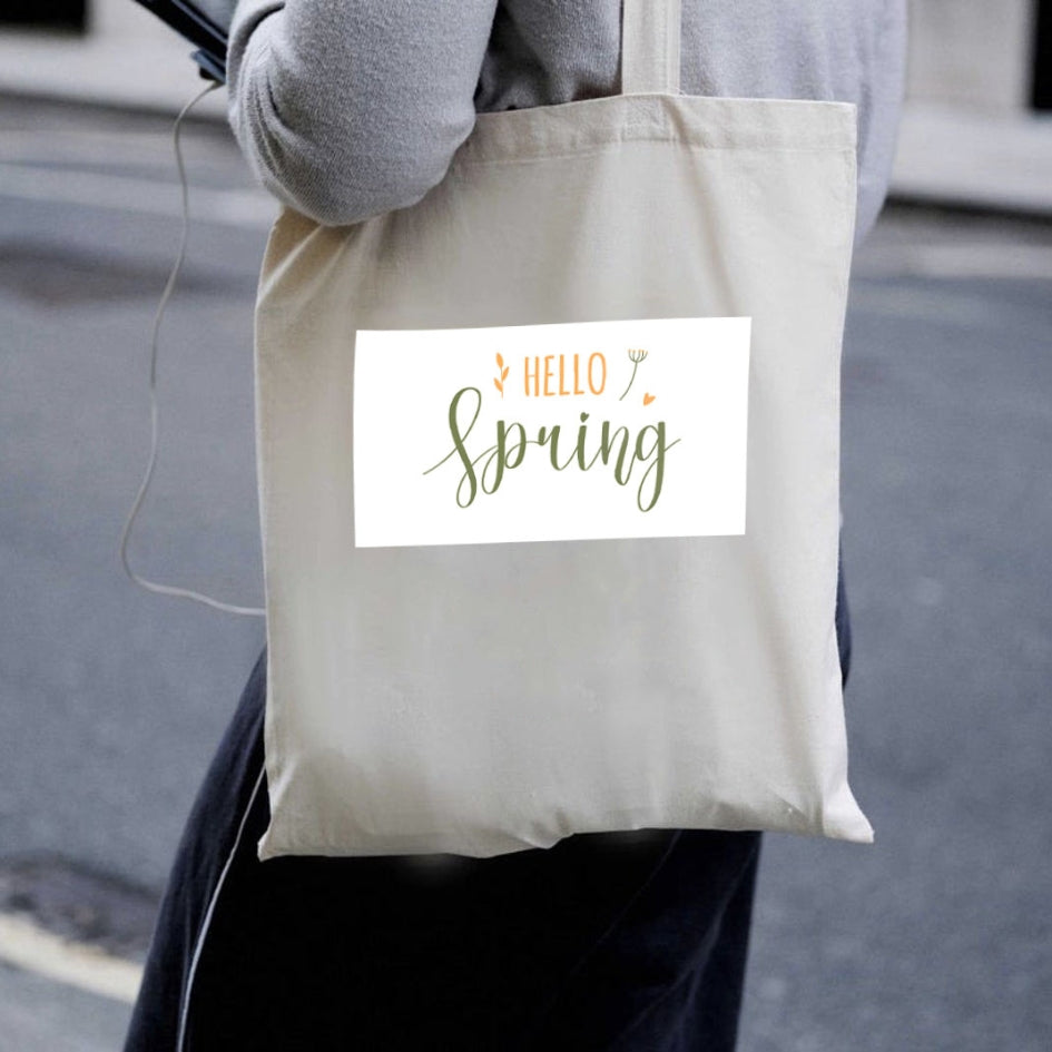 Prírodná plátená taška Kiskedves Hello Spring nesená cez rameno, so zeleno-oranžovým nápisom hello spring, v uliciach mesta.