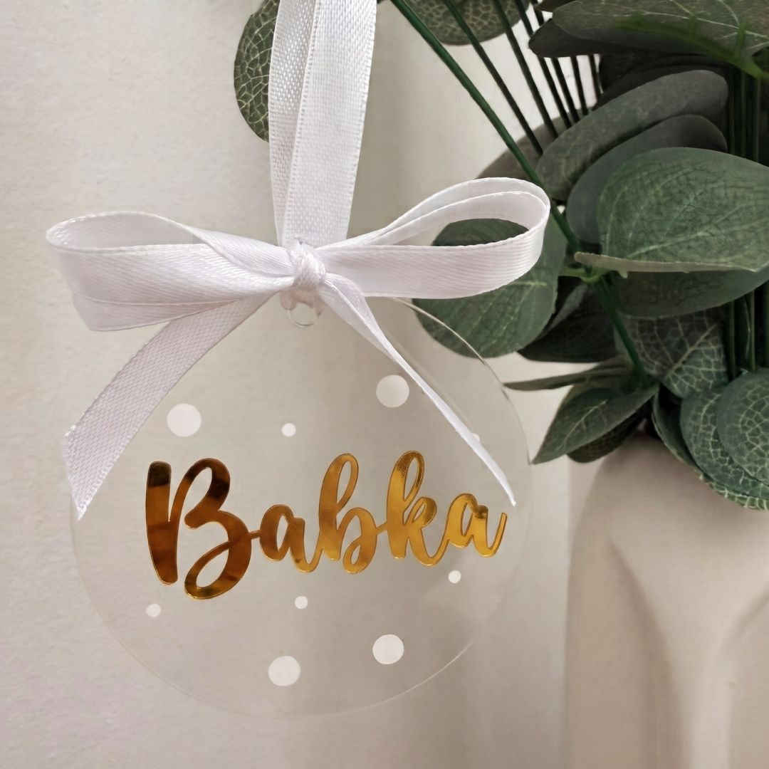 Priehľadná akrylová vianočná ozdoba so zlatým nápisom „Babka“, bielymi bodkami a saténovou mašľou
