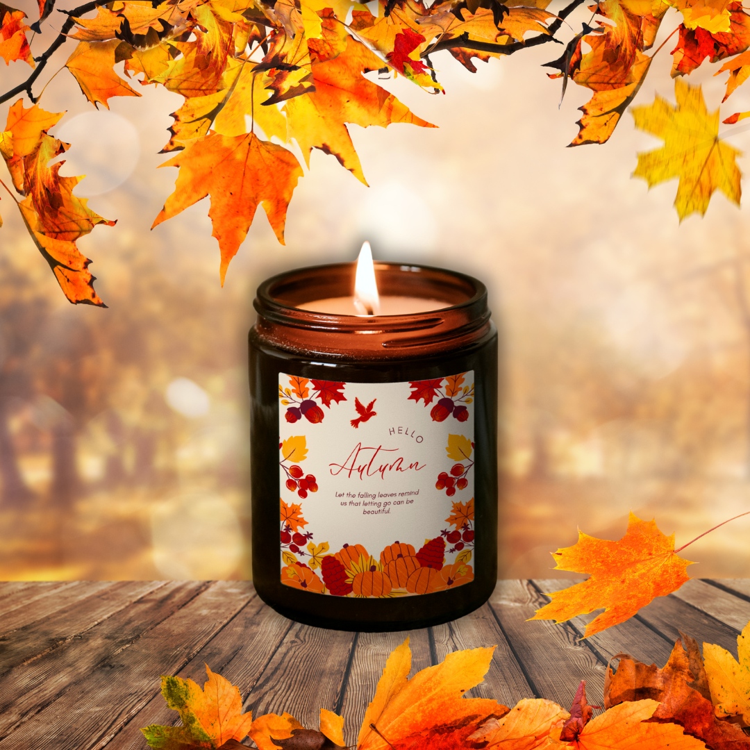 Horiaca sójová sviečka Pumpkin Spice v hnedom pohári so štítkom „Hello Autumn“, medzi jesenným lístím na drevenom stole