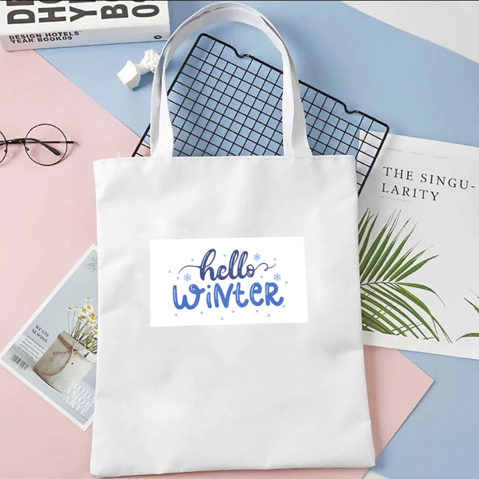 Plátená taška Kiskedves Hello Winter v bielej farbe, s modrým nápisom hello winter, na pastelovom pozadí vedľa časopisov a okuliarov.