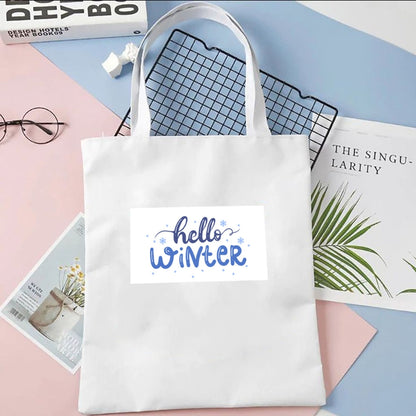 Plátená taška Kiskedves Hello Winter v bielej farbe, s modrým nápisom hello winter, na pastelovom pozadí vedľa časopisov a okuliarov.
