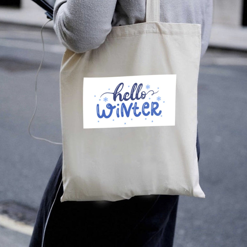 Prírodná plátená taška Kiskedves Hello Winter nesená cez rameno, s modrým nápisom hello winter, v uliciach mesta.