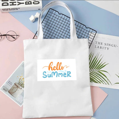 Plátená taška Kiskedves Hello Summer v bielej farbe, s oranžovo-modrým nápisom hello summer, na pastelovom pozadí vedľa časopisov.