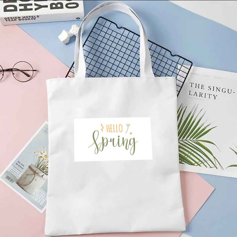 Plátená taška Kiskedves Hello Spring v bielej farbe, so zeleno-oranžovým nápisom hello spring, na pastelovom pozadí vedľa časopisov.