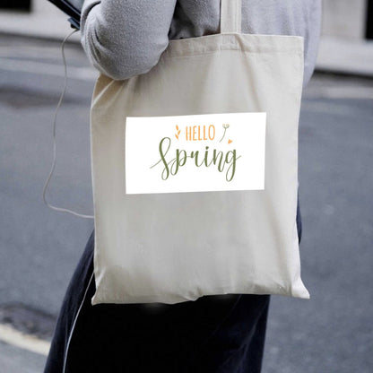 Prírodná plátená taška Kiskedves Hello Spring nesená cez rameno, so zeleno-oranžovým nápisom hello spring, v uliciach mesta.