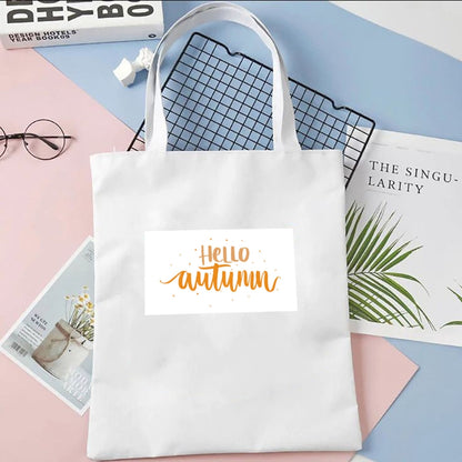 Plátená taška Kiskedves Hello Autumn v bielej farbe, s oranžovým nápisom hello autumn, na pastelovom pozadí vedľa časopisov.