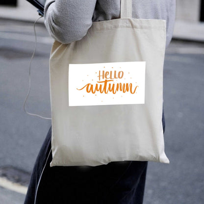Prírodná plátená taška Kiskedves Hello Autumn nesená cez rameno, s oranžovým nápisom hello autumn, v uliciach mesta.