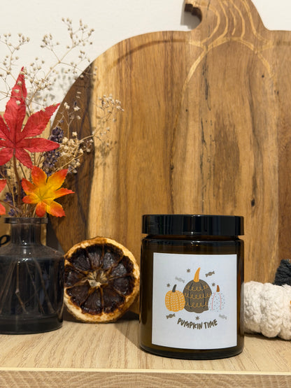 Sójová sviečka Pumpkin Spice v hnedom pohári so štítkom „Pumpkin time“, pred dreveným lopárom v tvare tekvice, vedľa váza so sušenými kvetmi, jesenné lístie a biela háčkovaná tekvica