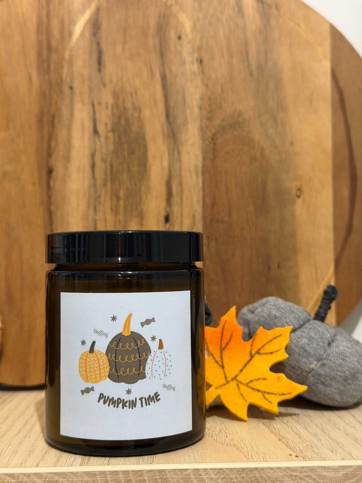 Sójová sviečka Pumpkin Spice v hnedom pohári so štítkom „Pumpkin time“, pred okrúhlym dreveným lopárom, vedľa žltý plstený javorový list a sivé látkové tekvice
