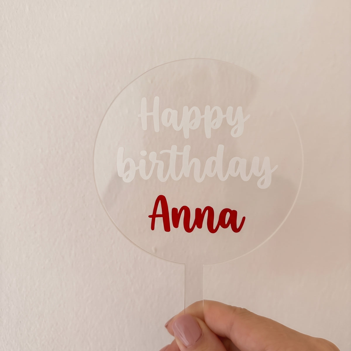 Priehľadný akrylový zápich do torty s bielym nápisom „Happy birthday“ a červeným menom „Anna“ v ruke