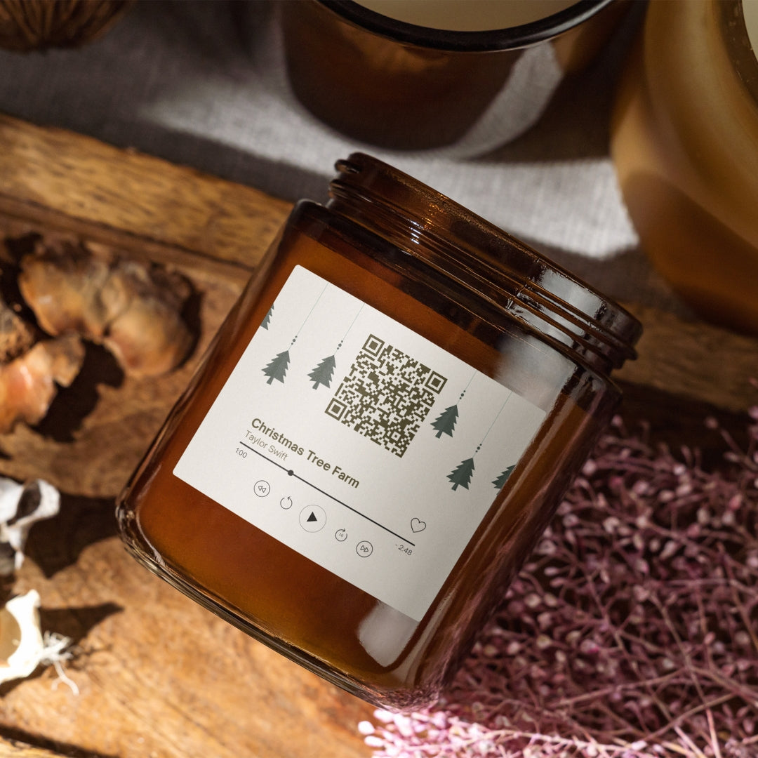 Vianočná sviečka „Melodická“ Kiskedves v hnedom pohári, na štítku QR kód a hudobná karta s piesňou Christmas Tree Farm