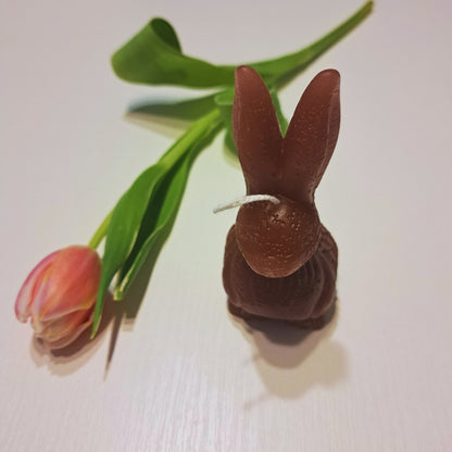 Hnedá sviečka Kiskedves v tvare zajačika s dlhými ušami na bielom drevenom stole, vedľa ružový tulipán.