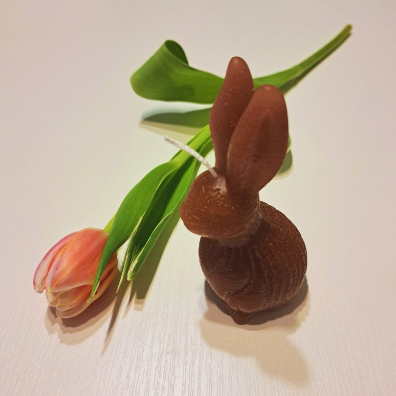 Hnedá sviečka zajačik od Kiskedves s okrúhlym telíčkom, na bielom drevenom povrchu s tulipánom.