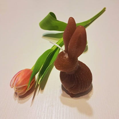 Hnedá sviečka zajačik od Kiskedves s okrúhlym telíčkom, na bielom drevenom povrchu s tulipánom.