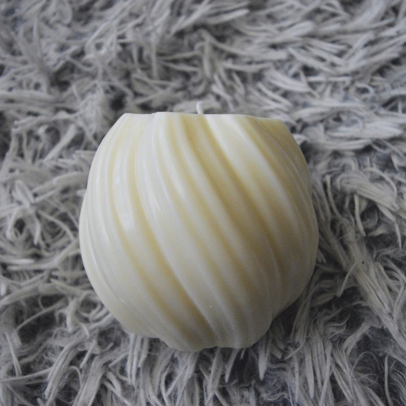 Sviečka Kiskedves Twist Glow: krémovobiela guľatá sviečka s točeným rebrovaním na sivom chlpatom koberci.