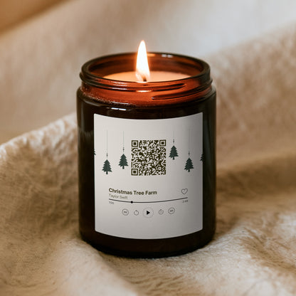 Horiaca vianočná sviečka „Melodická“ Kiskedves v hnedom pohári na mäkkej deke, na štítku QR kód a hudobná karta s piesňou Christmas Tree Farm
