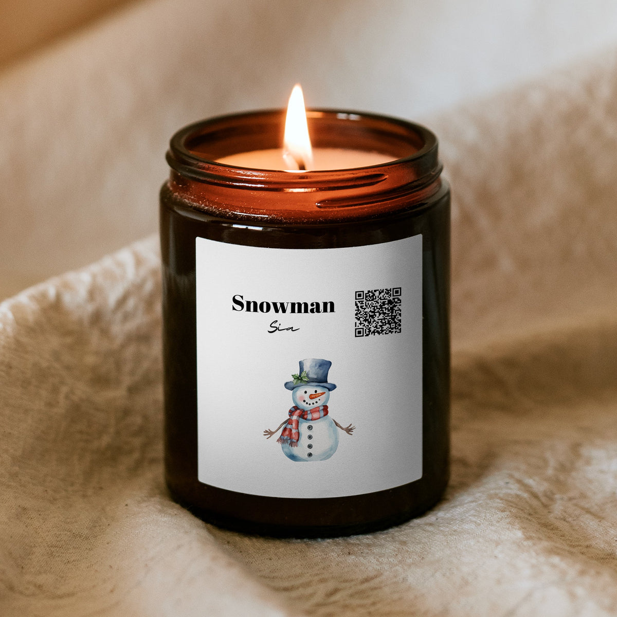 Horiaca vianočná sviečka „Melodická“ Kiskedves v hnedom pohári na mäkkej deke, na štítku QR kód, nápis Snowman a akvarelový snehuliak
